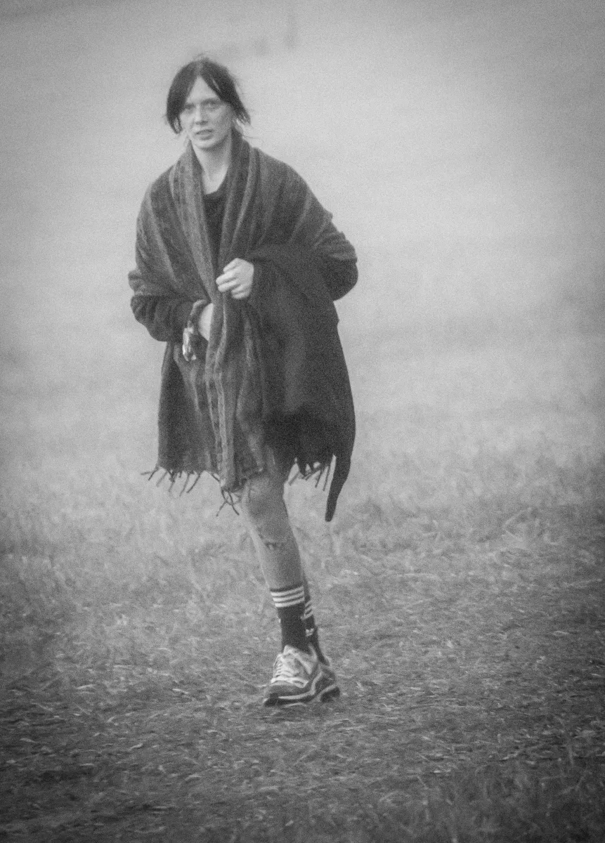 Woman wrapped in a dark shawl walking through fog across an open field